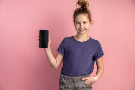 Portrait of a teen girl holding a smart phone with a black blank screen on a pink background. Girl isolated on blank background.の写真素材