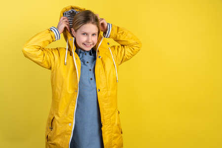 ?ute little girl puts on the hood of her raincoat. Isolated on yellow background beautiful little girl in a yellow raincoat.の写真素材