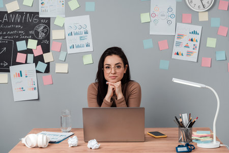 Pretty brunette with glasses sitting at the table with a laptop. On a gray background a lot of colorful stickers, charts, girl running.の写真素材