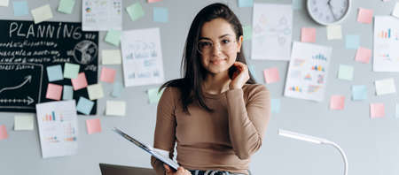 Beautiful brunette with papers in hands on a working background. She straightens her hair, smiles sweetly, looks at the camera.の写真素材