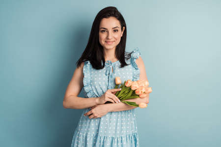 Young woman in a polka dot dress and flowers in her hands posing on a blue background. The concept of spring mood, copy space, place for textの写真素材