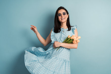 Beautiful, positive, young woman in a polka dot dress and flowers in her hands posing on a blue background. The concept of spring mood.の写真素材