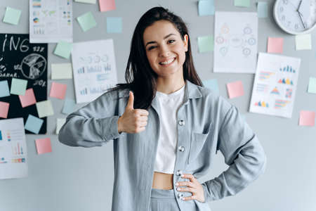 Happy businesswoman showing thumb up over gray background. The girl looks at the blue shirt, looks at the camera.の写真素材