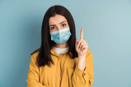 Beautiful brunette in a protected suit points her finger at a copy of the space on a blue level. Girl showing thumbs up, copy space, health concept.の写真素材