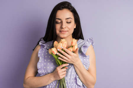 Charming brunette in a polka dot dress holds spring flowers on a purple background. The girl closed her eyes and sniffed the flowers.の写真素材
