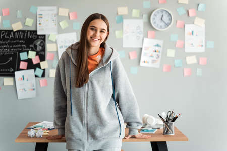 Smiling girl in a gray hooded sweater leaned against a desk in her office. Female student on a gray wall background with stickers, learning concept.の写真素材