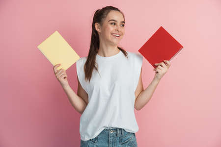 Cheerful teen woman smiling and showing laptops to camera while doing homework on pink background. Girl isolated on a blank wall background.の写真素材