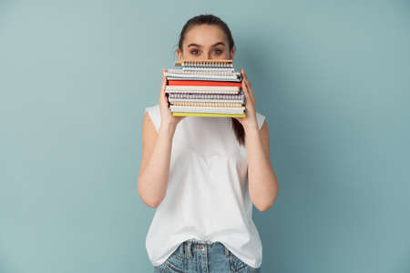 Concerned girl studying holding notebooks and books. Isolated on a blue background, the student covers half of her face with her notes.の写真素材