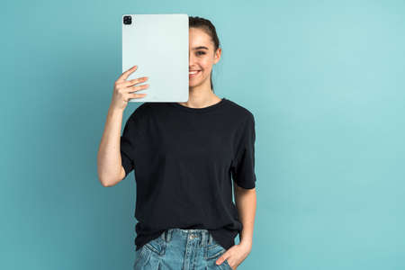 Cute girl covers half of the face with a tablet. Girl in a black T-shirt isolated on a blue backgroundの写真素材