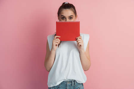 Portrait of a beautiful young woman wearing white t-shirt and blue jeans standing isolated over pink background. Beautiful girl covers her face with a notebook.の写真素材