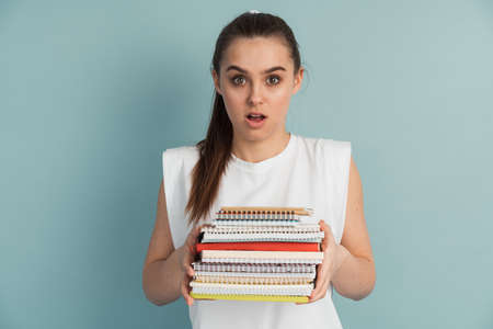 Cute female student with a mountain of notebooks and books in her hands on a blue background. Girl on the background of an empty wall, she looks surprised.の写真素材
