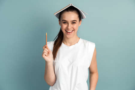 Cheerful, funny girl with a notebook on his head in his pencil. The girl has fun on a background of a blue wall.の写真素材