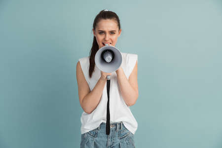 Young girl in white t-shirt isolated on blue background screaming in megaphone. Advertisement concept. Mock up copy space.の写真素材