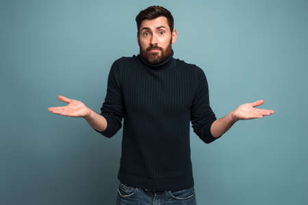 I don't know. Portrait of confused handsome bearded young man standing with raised arms and looking at camera with answer. Indoor studio shot, isolated on blue backgroundの写真素材