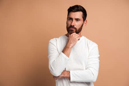 Portrait of pensive handsome bearded young man standing, touching his face, looking aside away and thinking about something. Indoor studio shot, isolated on beige backgroundの写真素材