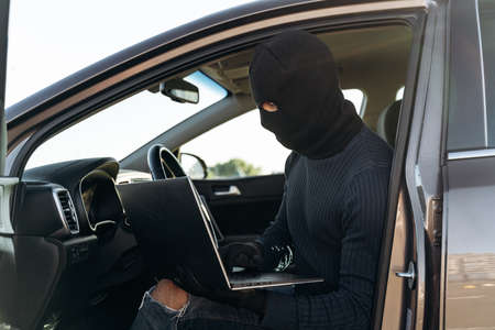 Masked thief in a balaclava stealing laptop from car and looking at the screen while sitting inside. Criminal concept. Stock photoの写真素材