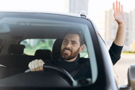 Saying hello. Handsome young smiling businessman sitting in new car and waving to someone while driving the car with pleasure emotionsの写真素材