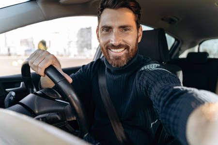Great selfie. Handsome bearded man holding camera and making selfie while sitting in the car at the driver seat and looking at the camera with pleasure smileの写真素材
