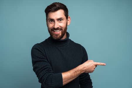 Advertise here. Positive bearded man pointing finger away paying your attention at empty space for advertisement, looking at camera with toothy smile. Indoor studio shot isolated on blue backgroundの写真素材