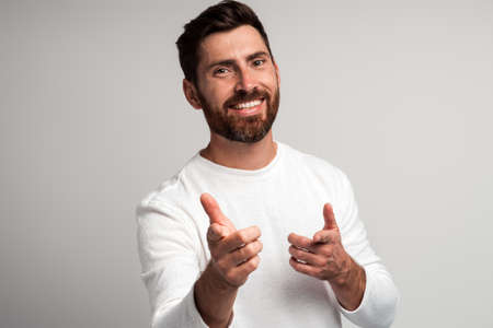 Hey you. Portrait of happy bearded man in white shirt smiling and pointing to the camera against light gray background. Studio shotの写真素材