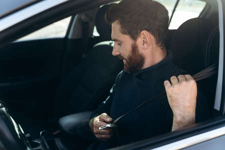 Side view of the caucasian young bearded man in a car fastening his seat belts before the driving. Confident guy riding at the work. Stock photoの写真素材