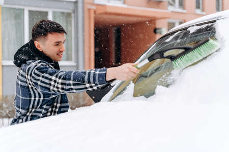 Man shoveling and removing snow from his car during a snow storm. Caucasian male standing with special brush and removing snow from the windowの写真素材
