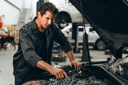 Caucasian male mechanic repairs car in garage. Man attentively looking at the car. Car maintenance and auto service garage conceptの写真素材