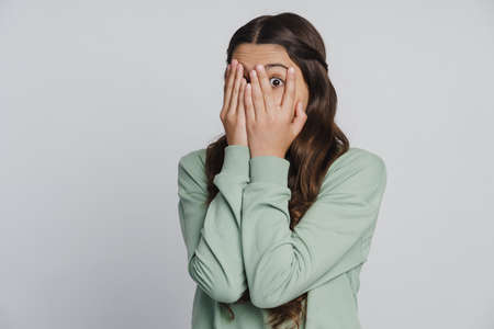 Beautiful girl closes her eyes with her hands, looking through her fingers. The girl spies with fear through her fingersの写真素材