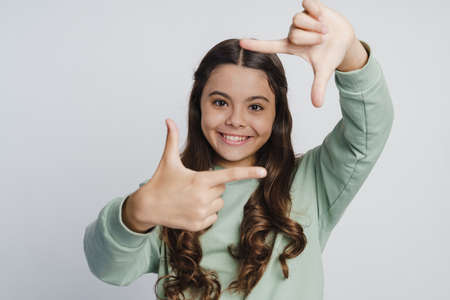 Smiling, positive girl posing on a white wall background. The child shows a frame with his fingers.