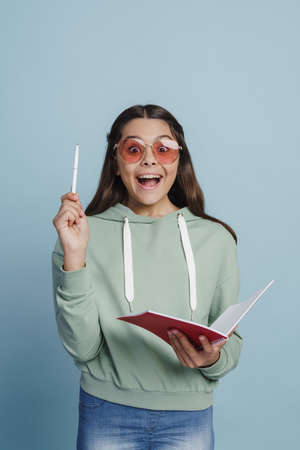 Attractive teenage girl has an idea, raised the pen up, holding a notebook. Cute little girl posing on a blue backgroundの写真素材