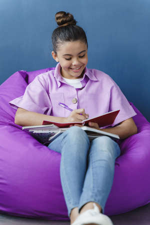 Attractive, positive teenage girl sitting in a comfortable pouf, studying with a smile. Little girl...