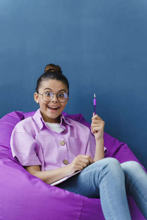 Cheerful, positive teenage girl with a smile has a great idea, lifted the pen up, sitting in a comfortable purple ottoman. Little girl on a blue backgroundの写真素材