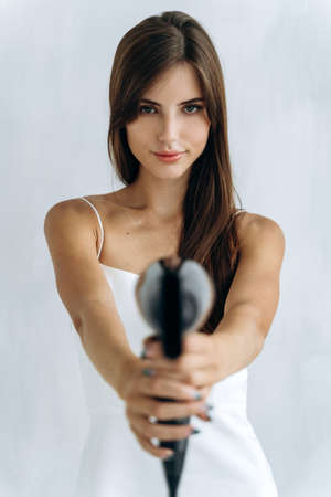 At gunpoint. Beautiful happy woman drying healthy long straight hair using hair dryer. Portrait of attractive girl with brunette hair holding hairdryer like a gun. Hairstyle and hairdressing conceptの写真素材
