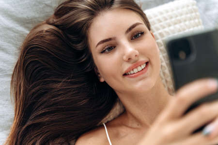 Delighted woman using smartphone in bed. Young smiling woman laying on the bed at home and texting at her smartphone, she is social networking and chatting. Stock photoの写真素材