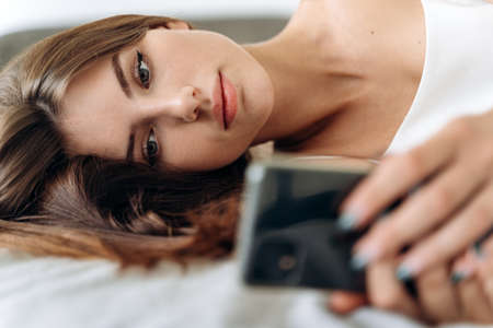 Romantic woman looking at mobile phone while laying on her bed. Brunette young woman using cellphone at home. Beautiful girl surfing the net on smartphone and relaxingの写真素材