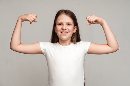 Look at my muscle, I'm strong. Portrait of adorable little girl in T-shirt showing her biceps and strength, feeling powerful and self-confident. Indoor studio shotの写真素材