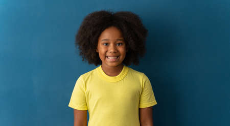 Portrait of cheerful little girl with curly hair in T-shirt smiling funny and carefree, showing front teeth. Healthy happy child, positive emotions concept. Indoor studio shot isolated on blue backgroundの写真素材