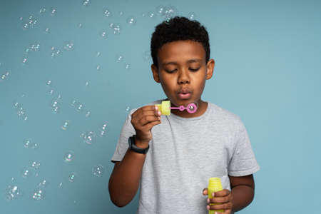 Attractive multiracial child boy in white T shirt standing and blowing soap bubbles over blue background. Happy childhood conceptの写真素材