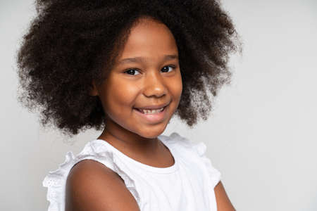 Closeup portrait of amazing cheerful little girl with curly hair in white T-shirt looking at camera with happy carefree smile. Indoor studio shot isolated on white backgroundの写真素材