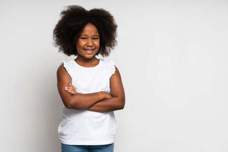 Portrait of funny little girl in T-shirt thinking over her idea and smiling at camera, while posing arms crossed. Indoor studio shot isolated on white backgroundの写真素材