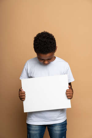 Vertical view of the beautiful multiracial boy holding a blank white sheet of paper and looking at it with interest. Space for text, advertisingの写真素材