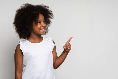 Look, advertise here. Portrait of cute cheerful little girl with curly hair in white T-shirt pointing to empty place on background. Preschooler showing copy space for promotional ad. Indoor studio shotの写真素材