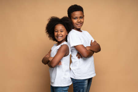 Waist up portrait view of the lovely multiracial brother and sister standing back to back to each other and smiling while posing at the studio over the beige wallの写真素材