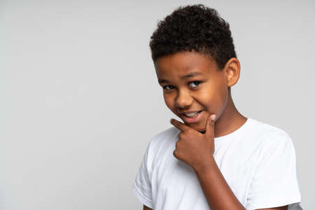 Portrait of thoughtful beautiful brunette young boy with black hair in white shirt standing, touching his chin, looking at the camera and thinking. Indoor studio shot isolated on white backgroundの写真素材