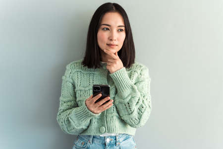 Portrait of romantic brunette woman using cellphone and pondering, reading good news message, enjoying mobile application. Indoor studio shot isolated on grey backgroundの写真素材