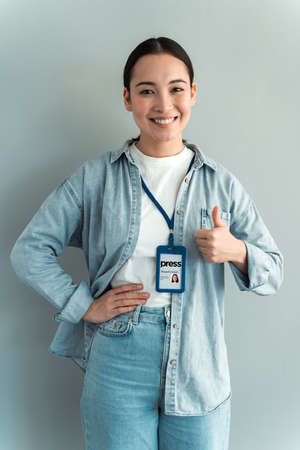 Work are done! Portrait of young woman with badge wearing jeans shirt, standing and showing thumbs up and looking at camera. Grey background, indoor studio shot isolatedの写真素材