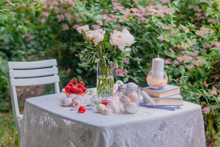Pink peonies in a glass vase and books in lilac covers, sweets, marshmallows and ripe strawberries in a bowl on a white table in the backyard garden.Photo of a beautiful decor for a photo shoot and decoration of a country houseの写真素材