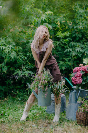 Beautiful girl with long blonde hair holds a watering can with flowers in a blooming gardenの写真素材