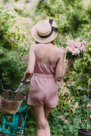 A beautiful stylish girl in a straw hat and a pale pink powdery jumpsuit walks with a mint-colored Bicycle in the garden of a country house. She holds a bouquet of fresh pink peonies in her hands. Stands with his back to the cameraの写真素材
