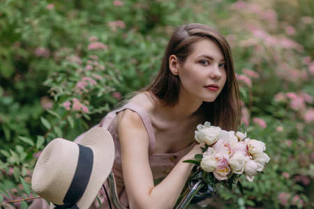 Portrait of beautiful girl with peony bouquet walks with bicycle in gardenの写真素材